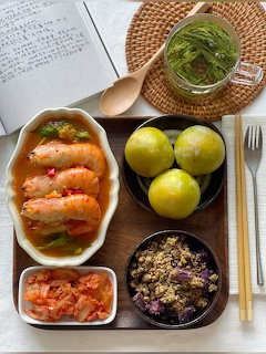 Overhead view of Korean food on a wooden tray with shrimp, kimchi, limes, grain bowl, and herb jar