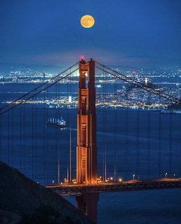 Golden Gate Bridge tower at dusk with a full moon above and city lights across the bay