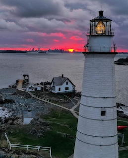 White lighthouse tower beside small white house on rocky shoreline at sunset with pink clouds and distant cityscape