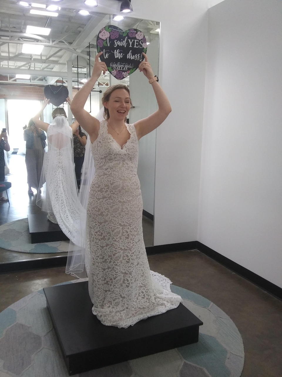Smiling bride in a lace wedding dress holding a sign in a bridal shop