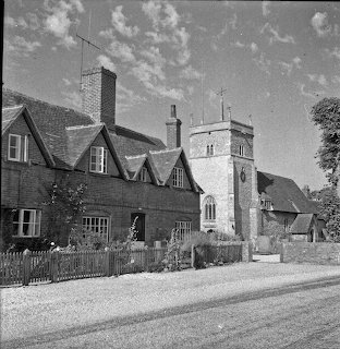 Black-and-white village street with cottages and a stone church tower under a cloudy sky