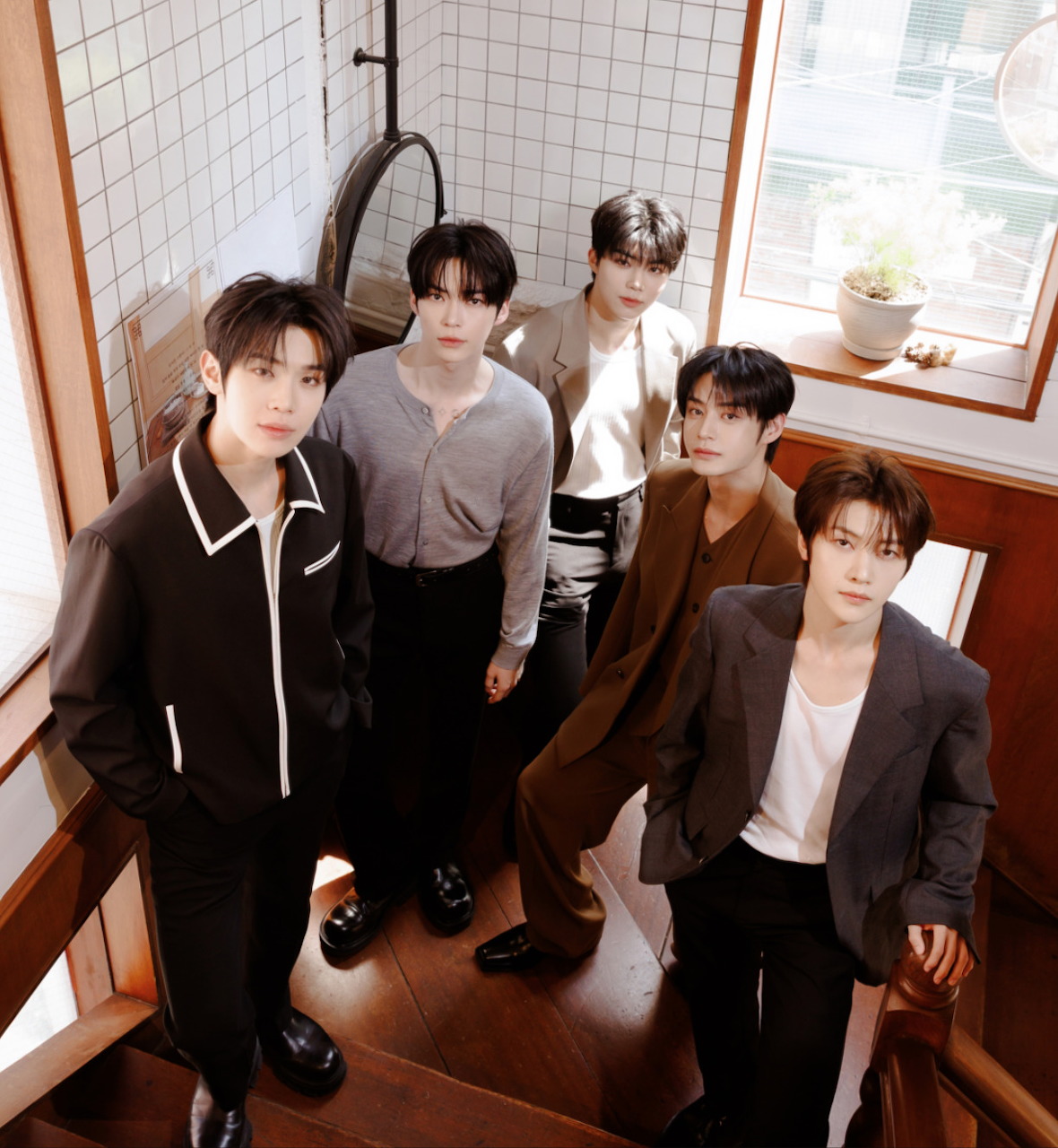 Five young men in dark casual outfits standing together in a sunlit room with wooden floor and tiled walls