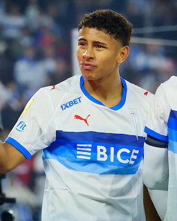 Young soccer player in a white and blue jersey with a blurred stadium crowd behind him.