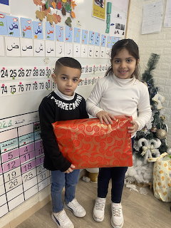 Boy and girl holding red ornate poster in classroom with Arabic alphabet chart behind them