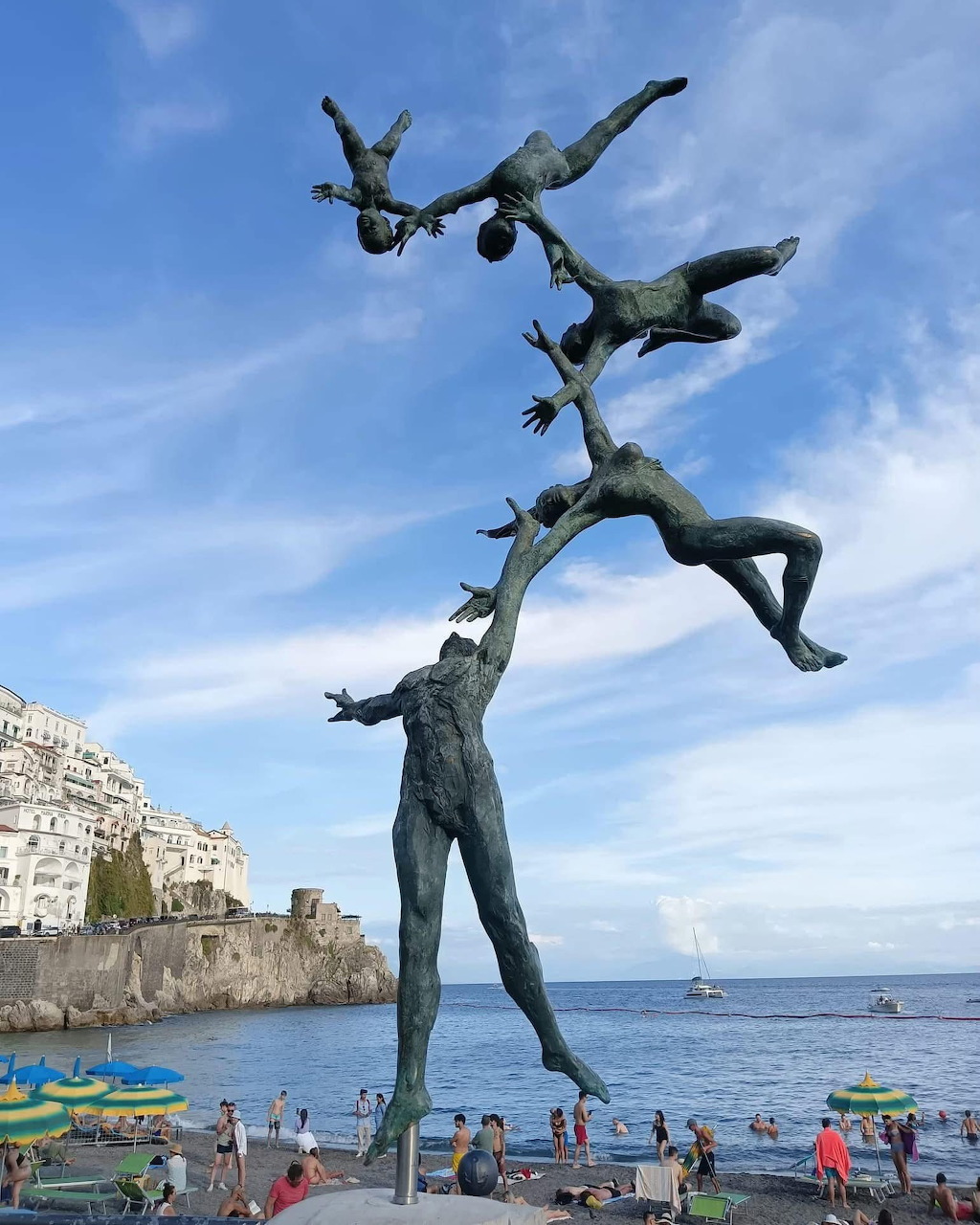 Bronze sculpture of stacked human figures performing acrobatics against blue sky and coastal background in Amalfi