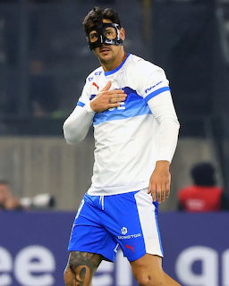 Soccer player in white jersey and blue shorts celebrating with hand on chest during night match