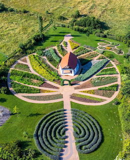Aerial view of a herb garden with curved paths, a small red-roofed building, and a circular maze