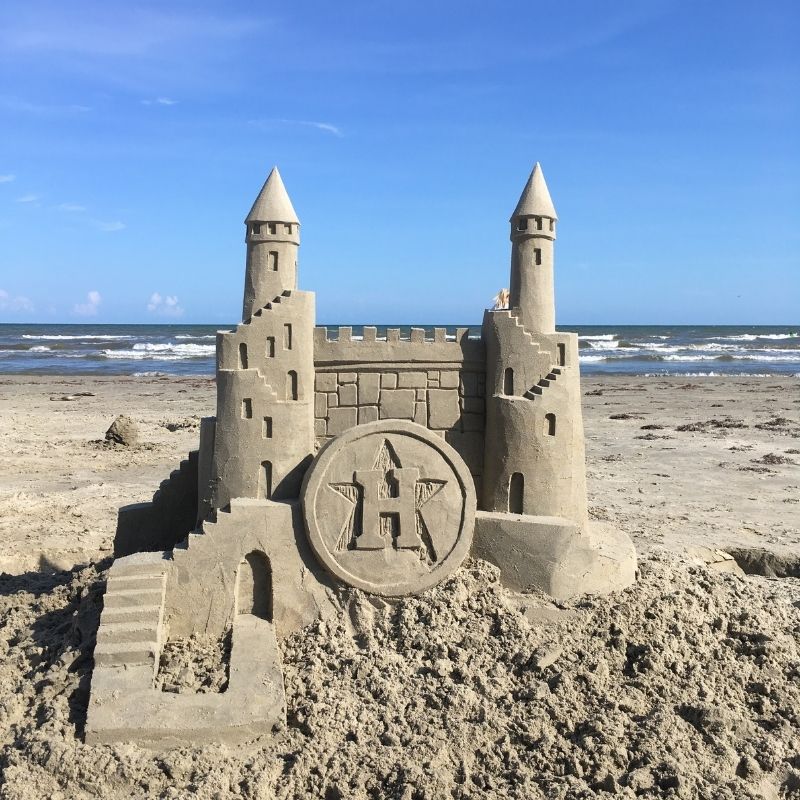 Sand castle with two towers and Houston Astros logo on beach with ocean backdrop