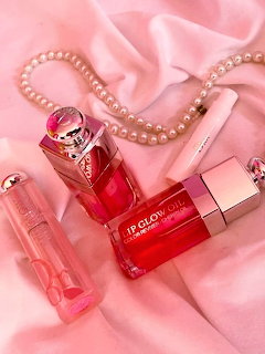 Flat lay of pink lip glow oil, lipstick, pearl necklace, and perfume bottle on pink satin fabric