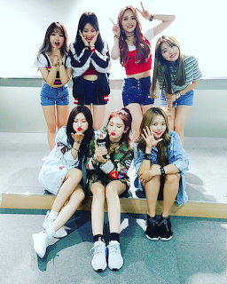Seven young women in colorful casual outfits posing together in a bright indoor setting