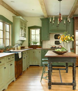 Country kitchen with green cabinets, wooden ceiling beams, rustic island table with yellow flowers and green fruit