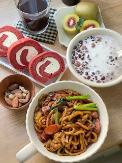 Overhead view of Korean food including noodles, red bean porridge, roll cake slices, nuts, and fruit on a wooden table