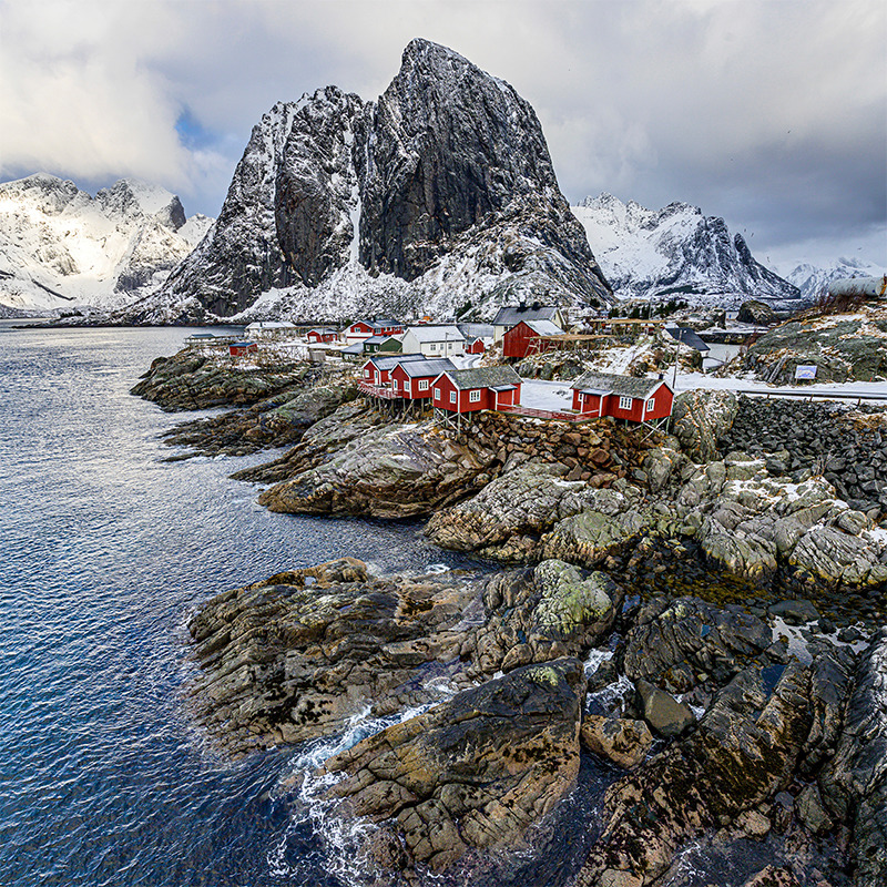 Red fishing cabins on rocky shore with snow-covered mountain peaks and fjord waters under cloudy sky