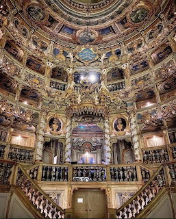 Elaborate baroque opera house with golden balconies, painted ceiling dome, crystal chandelier, and twin marble staircases