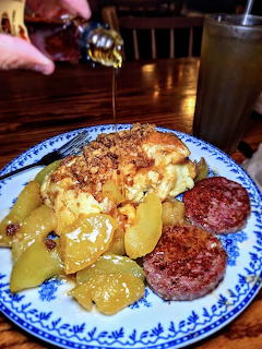 Breakfast plate with eggs, hash brown casserole, fried apples, and sausage with syrup being poured