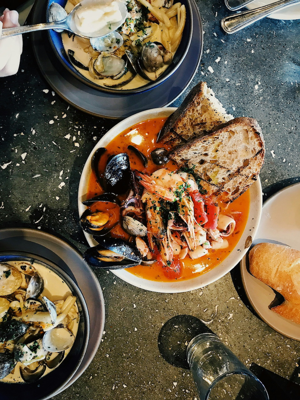 Bowl of cioppino stew with shrimp, mussels, clams and bread on speckled table with other bowls nearby