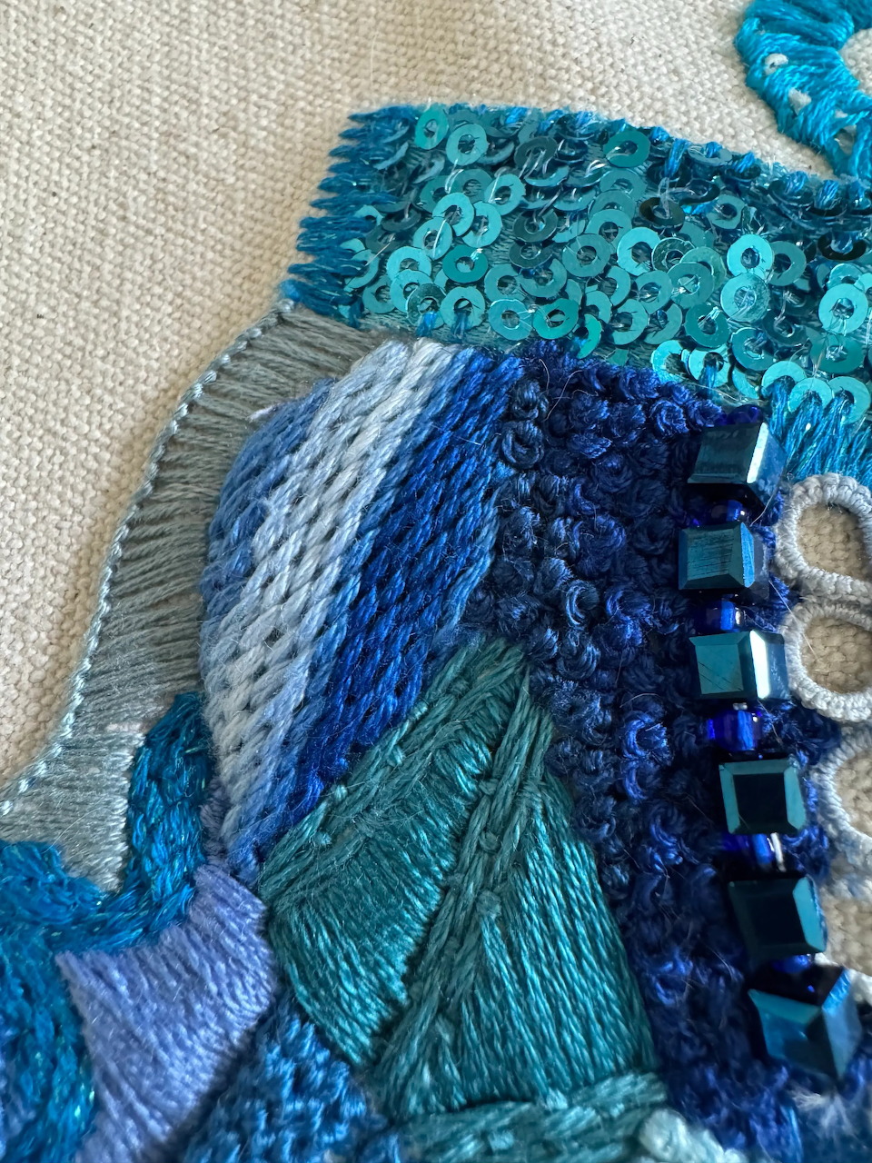 Close-up of blue embroidery sampler with various stitches, sequins, and beads on linen fabric