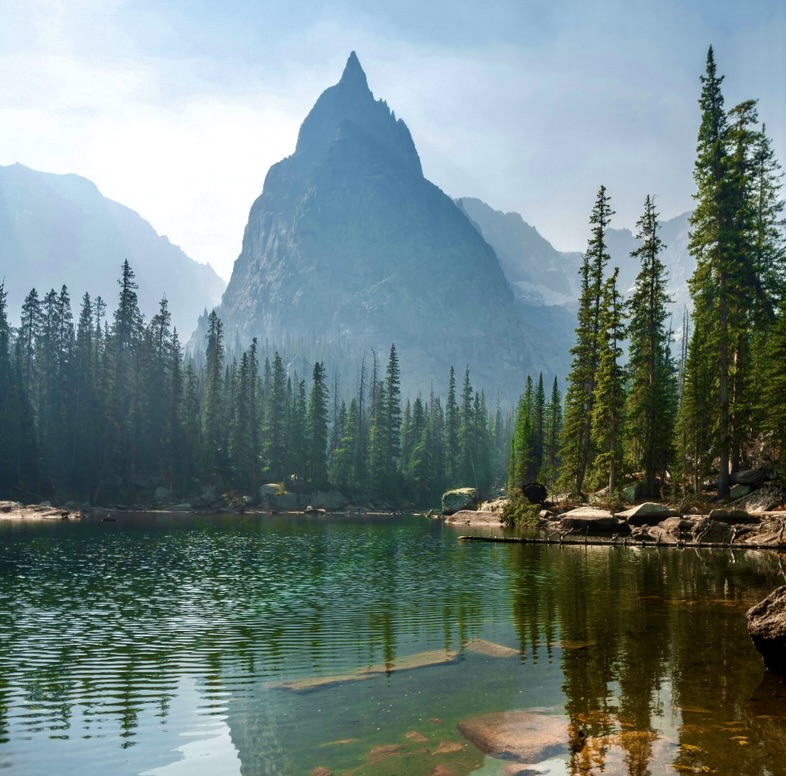 Sharp mountain peak towers over calm alpine lake surrounded by pine trees under hazy sky