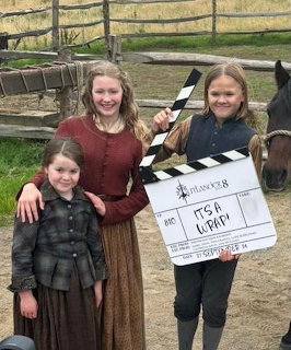 Three girls in historical costumes holding an Outlander Season 8 clapperboard on a farm set