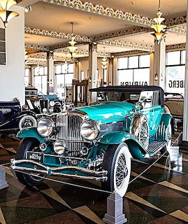 Teal vintage luxury car on display in an elegant automobile museum with checkered floor and chandeliers