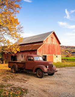 Rusty old truck parked in front of a red wooden barn with autumn trees and green hills in background