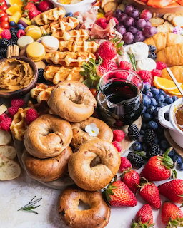 Overhead view of a brunch board with bagels, waffles, croissants, fresh fruit, and red wine glasses