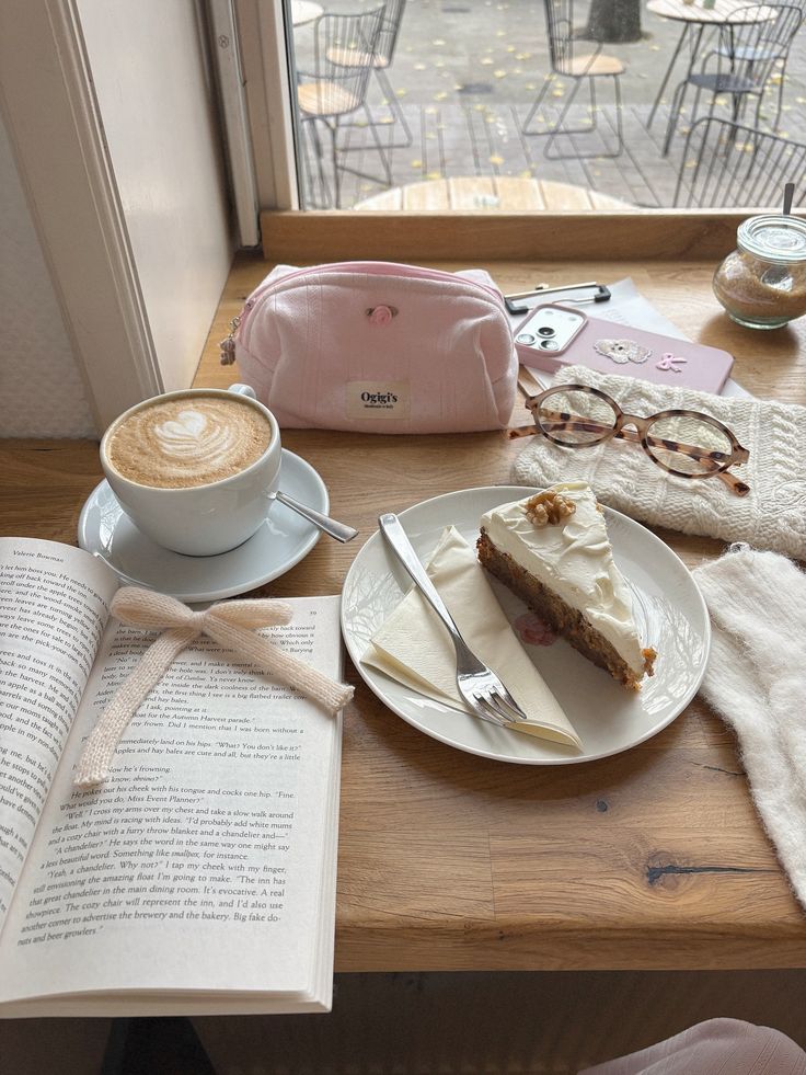 Cafe table with latte art coffee, cake slice, open book, glasses, and pink pouch near a window