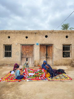 Women weaving a colorful traditional carpet outdoors in front of an earthen building