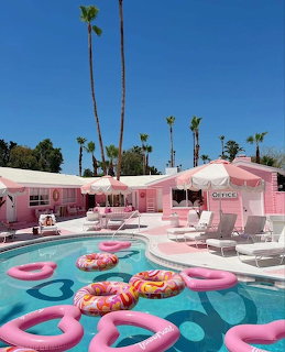 Pink pool with flamingo floats, donut inflatable, pink motel buildings, palm trees, and lounge chairs