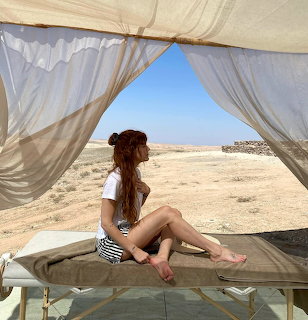 Woman in white shirt sitting on cushioned daybed under white canopy overlooking sandy desert landscape