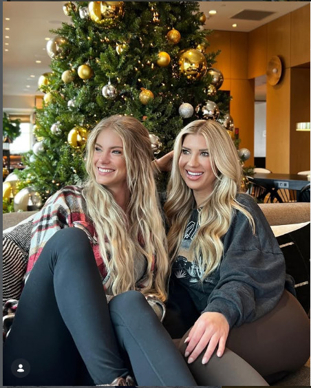 Two blonde women smiling together in front of a decorated Christmas tree indoors