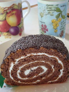 Chocolate Swiss roll cake with cream spiral on a plate and two floral mugs behind it