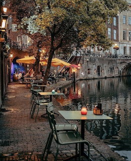 Outdoor café tables along a lit canal at dusk with trees and historic European buildings