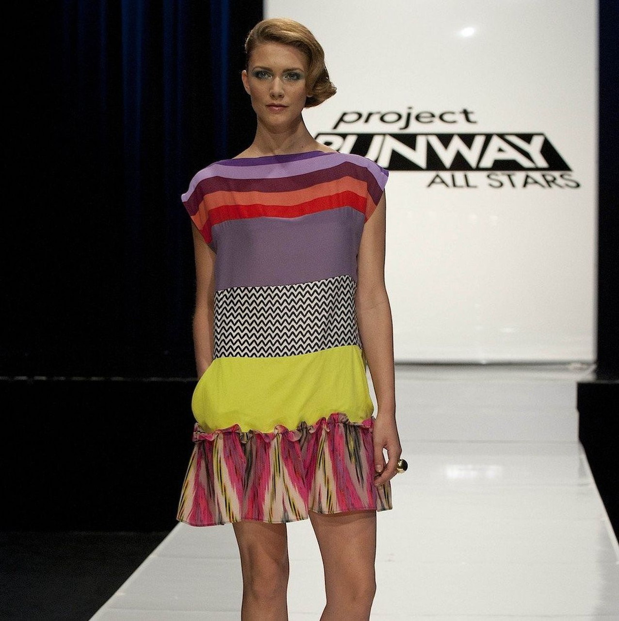 Model on a runway wearing a colorful striped dress with a Project Runway All Stars sign behind her