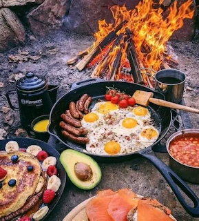 Breakfast cooking over campfire with eggs, sausages, pancakes and beans in cast iron pans