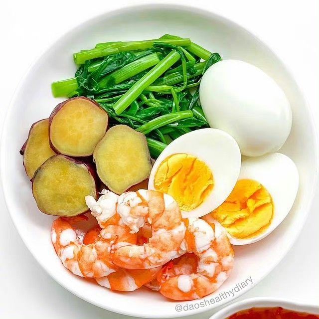 White bowl with boiled eggs, shrimp, sweet potato slices, and green vegetables on a white surface