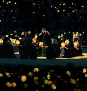 Singer on a dark stage with seated performers and glowing orb lights