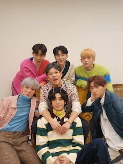 Seven K-pop boy group members posing together in colorful outfits against a beige wall