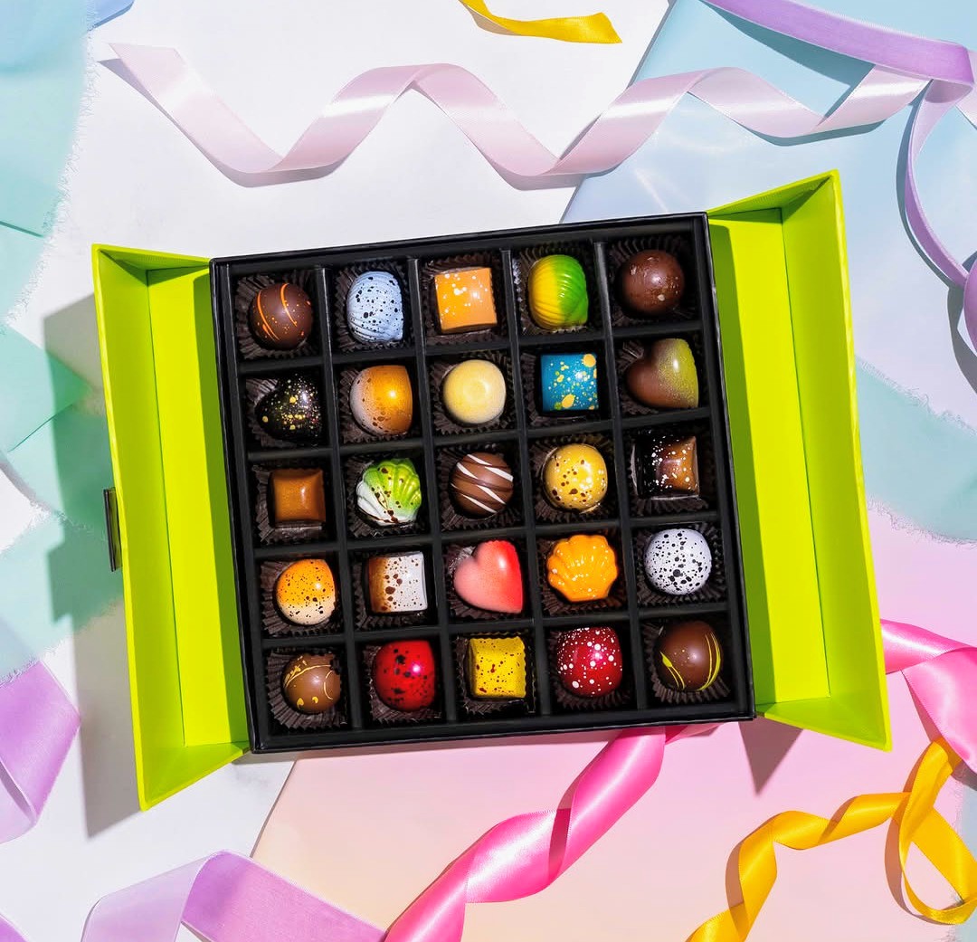 Open box of assorted chocolates with colorful ribbons on a pastel background