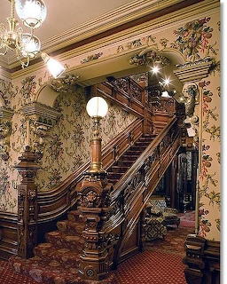 Ornate wooden staircase with intricate carvings in a Victorian mansion interior with floral wallpaper and chandelier