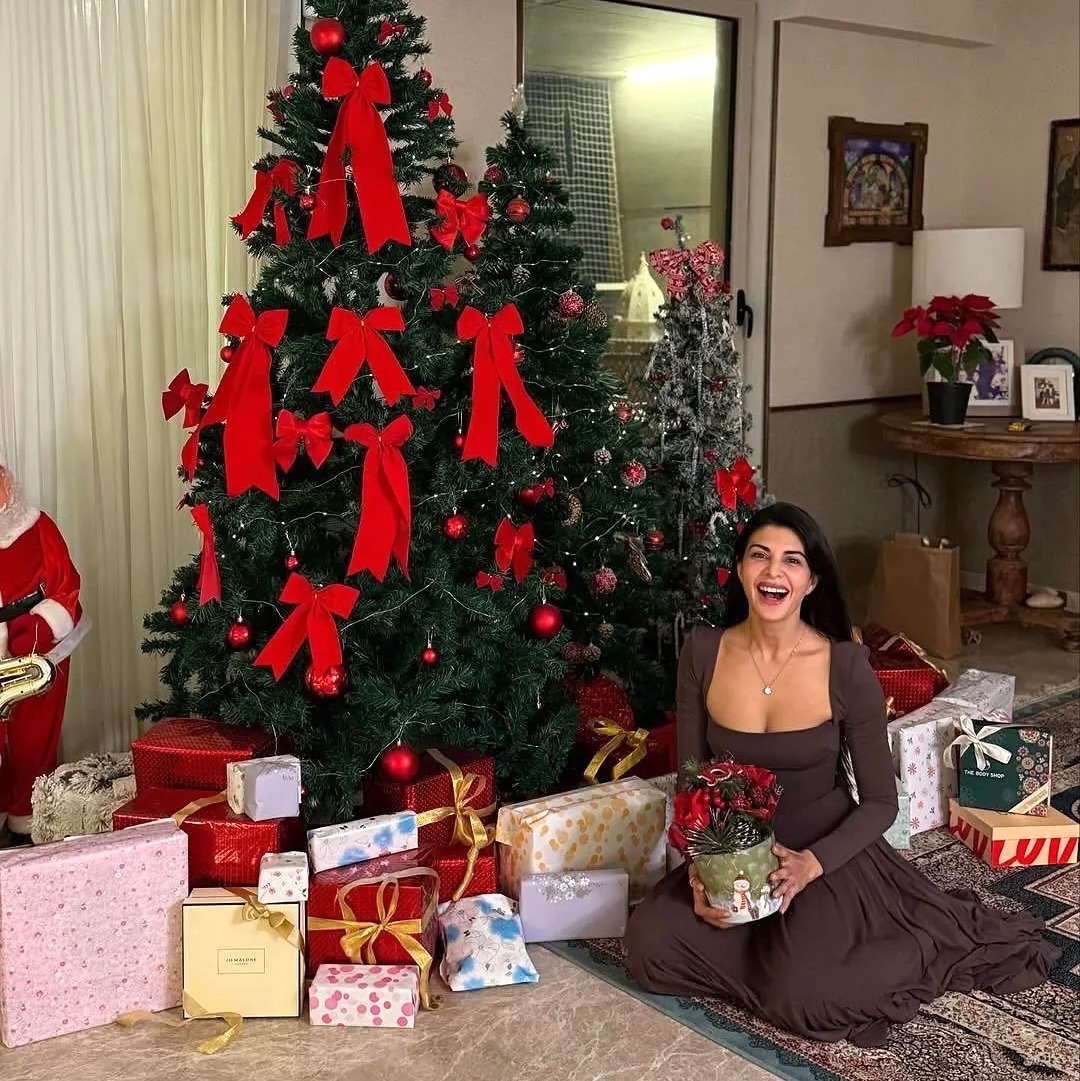 Woman in brown dress sitting by Christmas tree with red ribbons and presents in living room