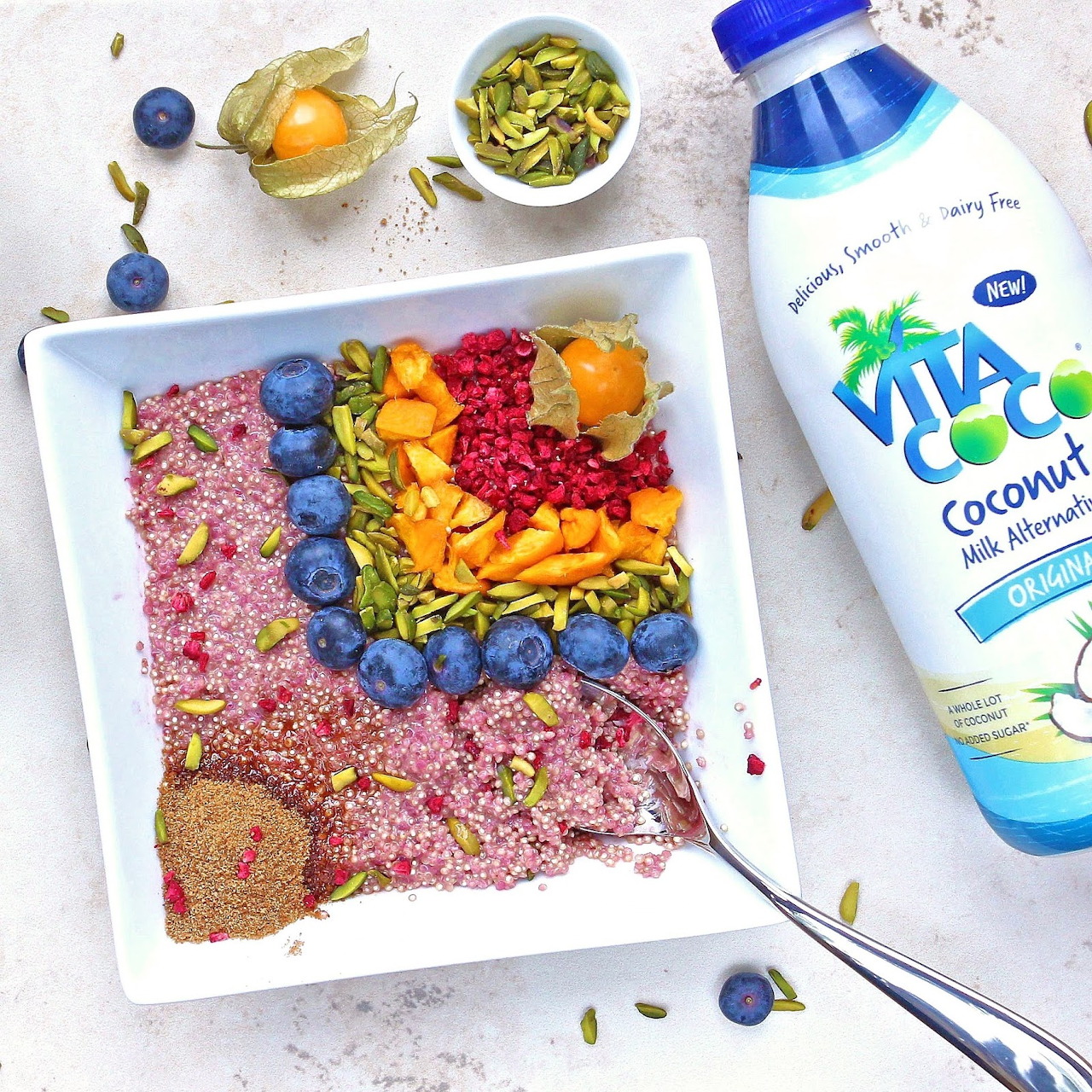 Rainbow breakfast bowl with colorful fruits, granola, seeds, and coconut water bottle on white marble surface