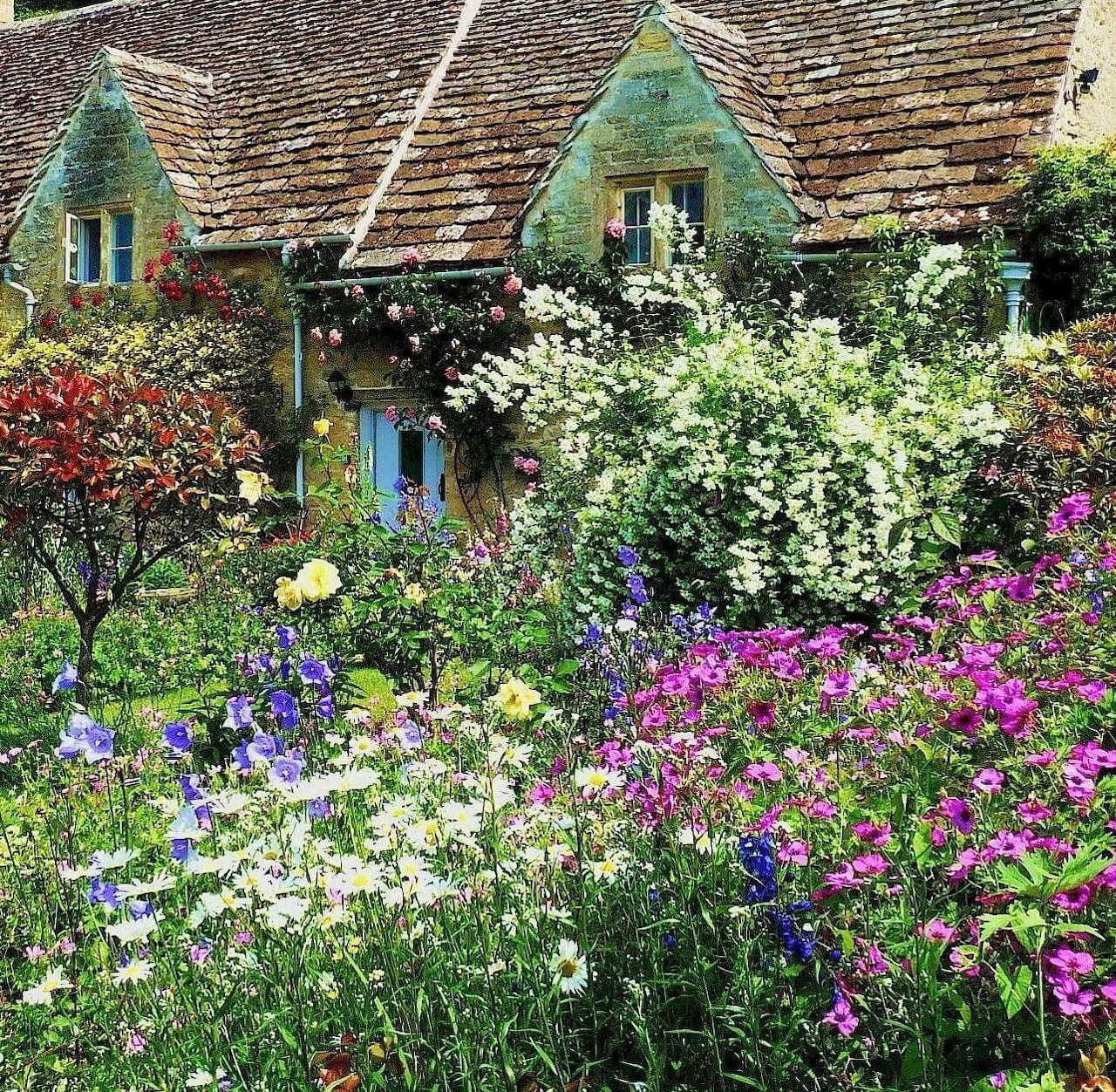 Stone cottage with dormer windows behind abundant colorful flower garden with pink, purple, and white blooms
