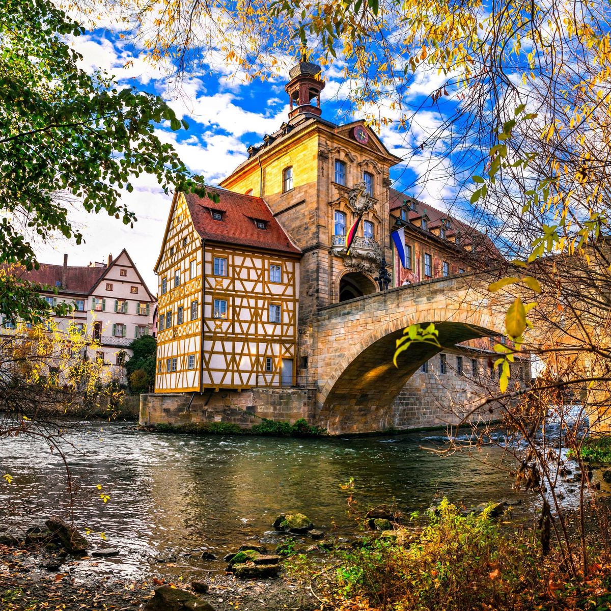 Historic half-timbered building on a stone bridge over a river in Bamberg, Germany, surrounded by autumn trees