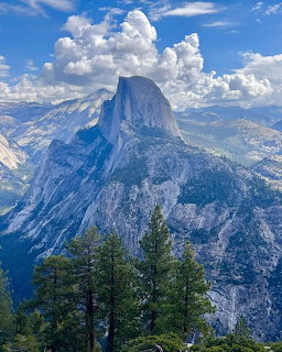Half Dome mountain above pine trees under a cloudy blue sky