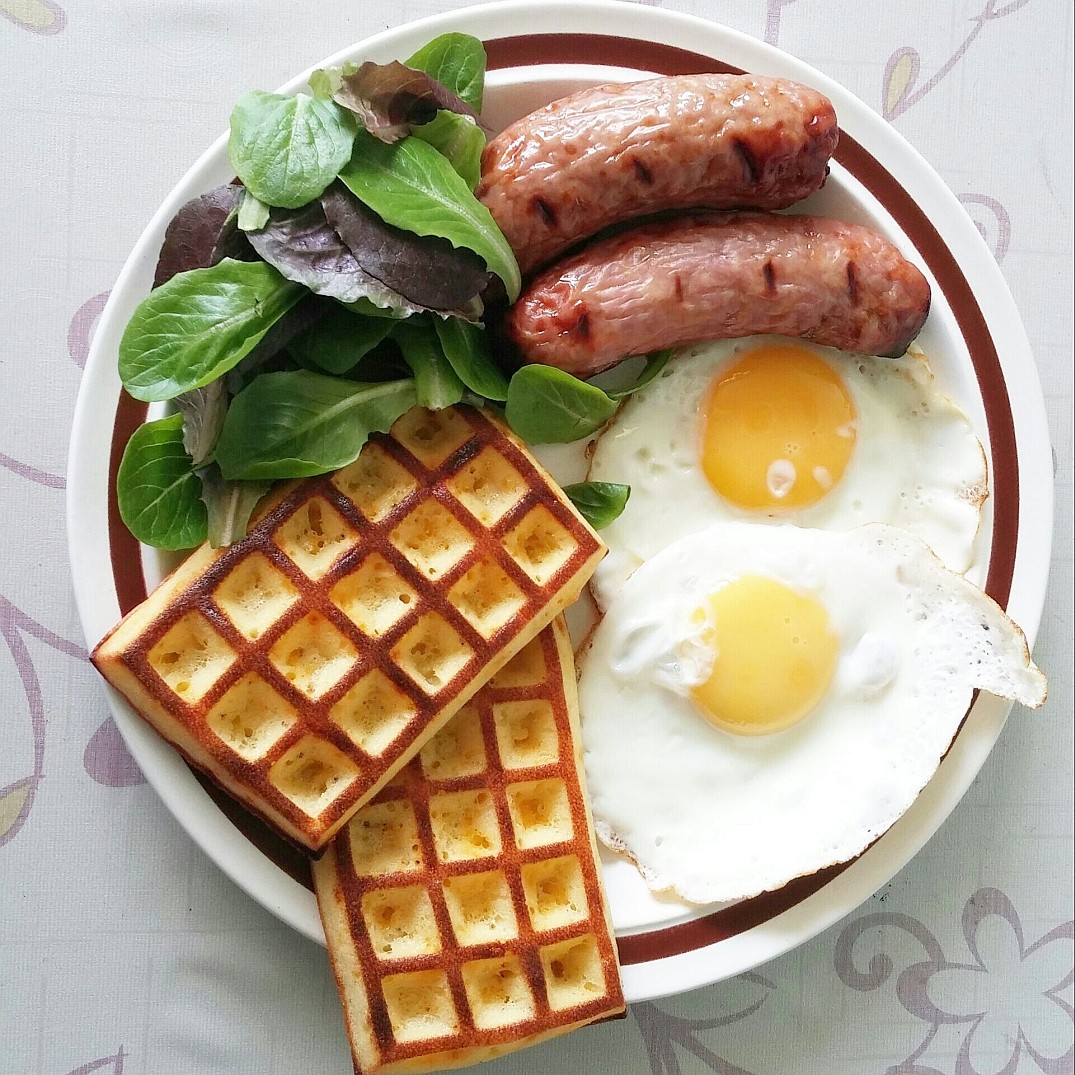 Overhead view of breakfast plate with waffles, fried eggs, sausages, and salad greens