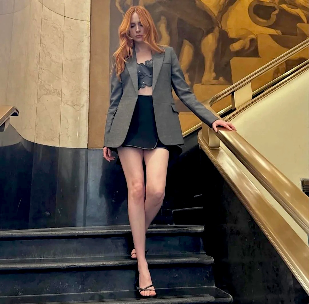 Red-haired woman in grey blazer and black mini skirt standing on marble staircase with gold railing