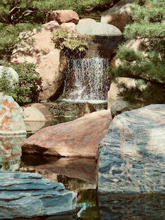 Small waterfall flowing over rocks into a reflective pond in a Japanese garden with pine trees