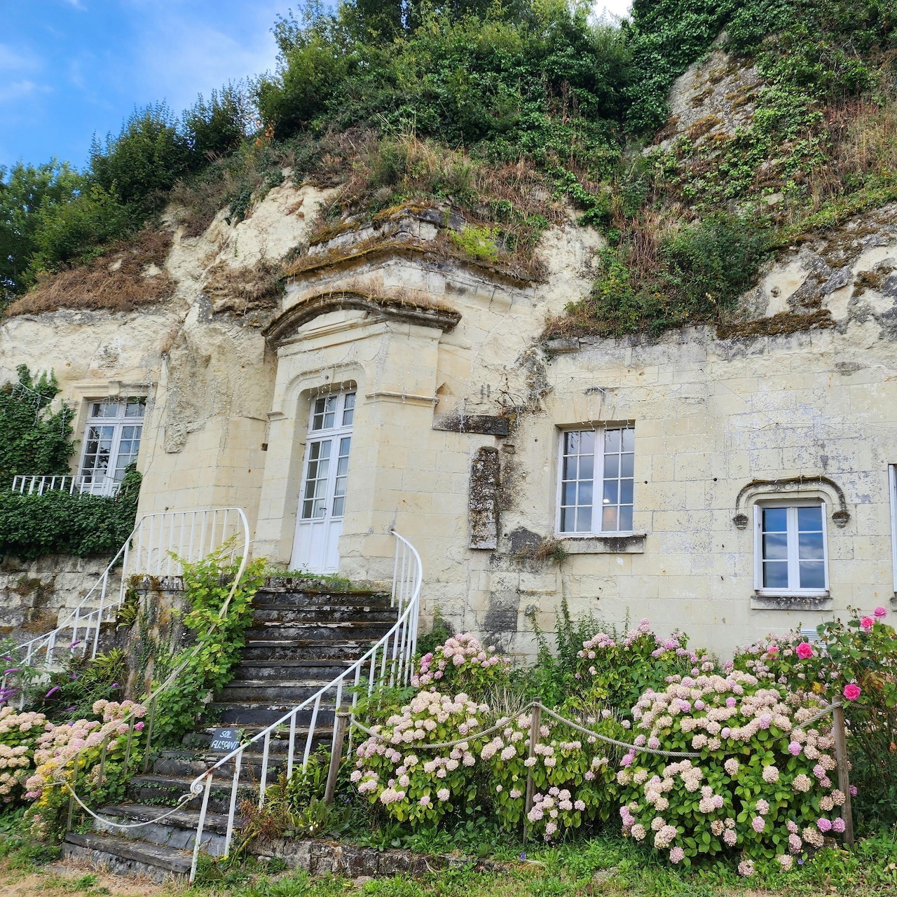 White-shuttered cave house built into rocky cliff with flowering garden and stone staircase