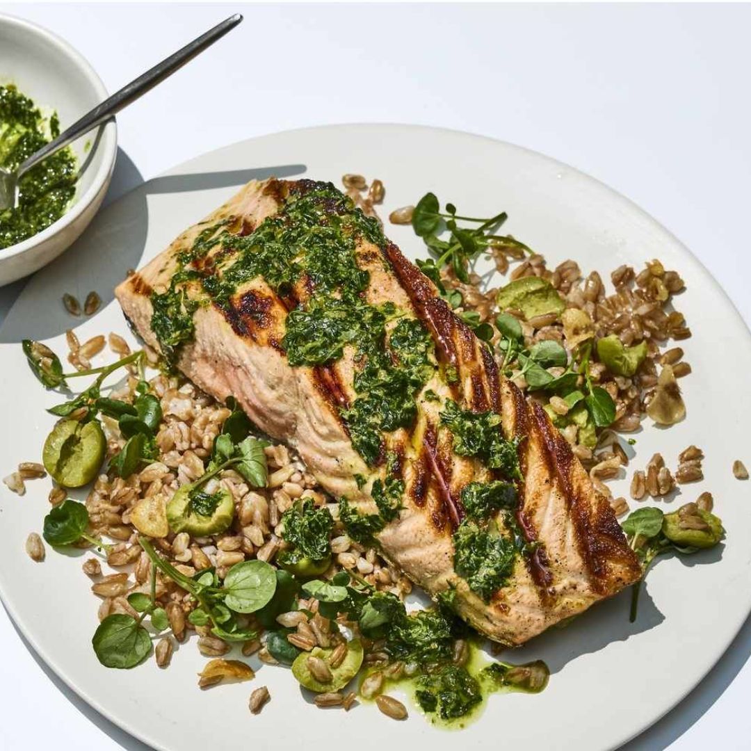 Grilled salmon on a white plate with grains, seeds, leafy greens, and green herb sauce